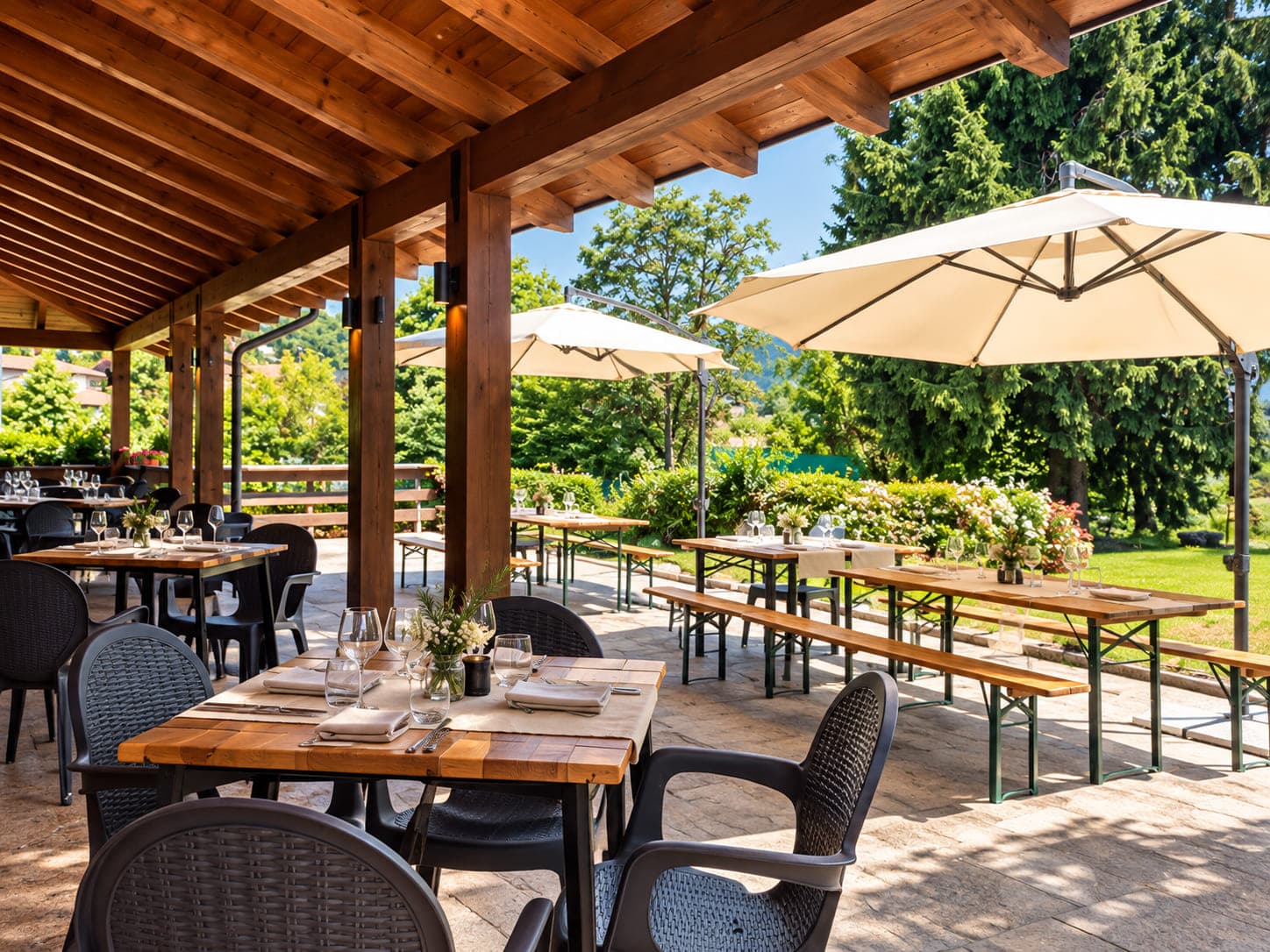 La terrazza coperta della trattoria: tavoli apparecchiati sotto il pergolato di legno, il giardino e le montagne sullo sfondo.
